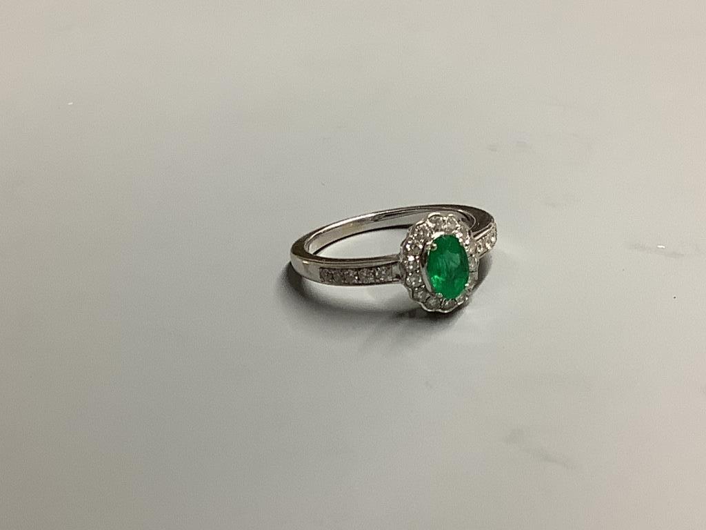 A modern 18ct white gold, emerald and diamond set oval cluster ring, with diamond set shoulders, size L, gross weight 3.4 grams.